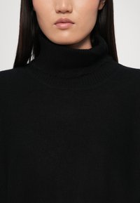 Schwarzer Rollkragenpullover aus einem weichen, strukturierten Stoff, mit einem gerippten Kragen und einer schlanken Silhouette. Nahaufnahme des Halsbereichs.