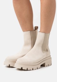 Beige Wildleder-Stiefeletten mit elastischen Seiteneinsätzen, runden Zehen und klobigen Gummisohlen mit tiefen Profilen für besseren Halt.