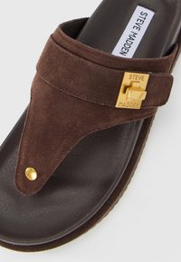 Brown suede thong sandal with gold-tone Steve Madden buckle and stud on black textured footbed.
