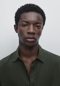 Portrait d'une personne avec des cheveux courts et texturés de couleur sombre, portant une chemise à boutons vert foncé avec col, sur un fond clair.