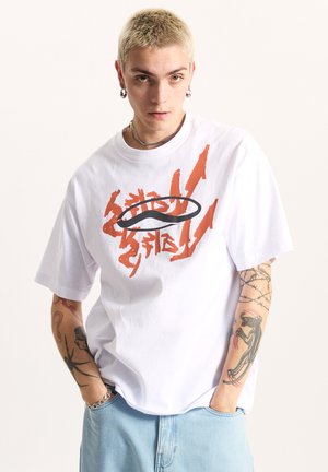 Hvid bomulds t-shirt med løs pasform, med orange grafisk tekst og et sort oval logo. Modellen bærer lyseblå denim shorts.