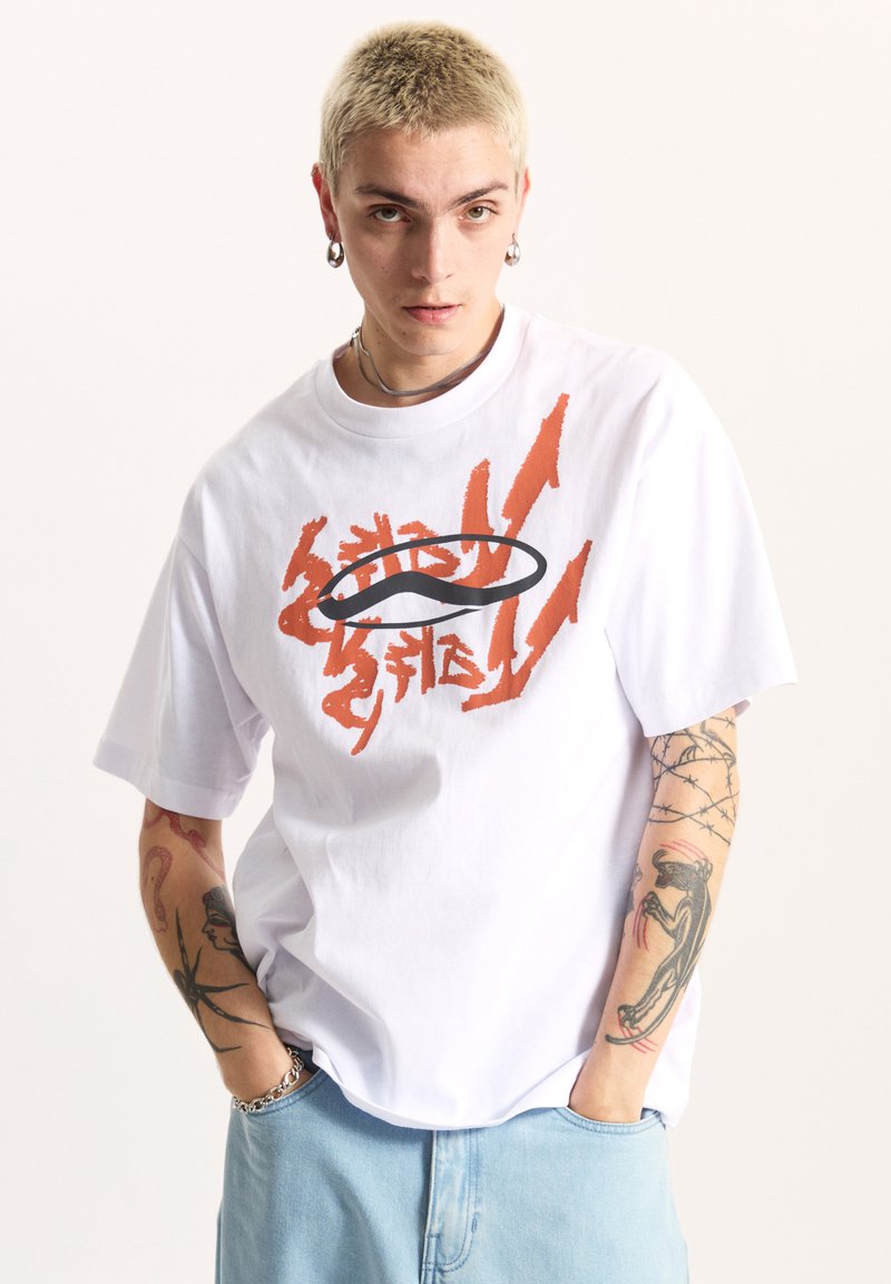 Camiseta blanca de algodón con un corte holgado, que presenta texto gráfico en naranja y un logo ovalado negro. El modelo lleva puesta unos pantalones cortos de mezclilla azul claro.