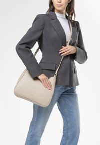 Borsa a tracolla in pelle beige con forma curvata e tracolla regolabile, esposta accanto a un blazer grigio e jeans blu.