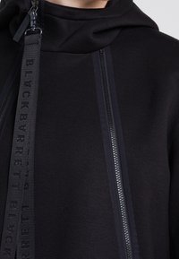 Svart hoodie i mjukt tyg, med två vertikala fickor med dragkedja och en framträdande märkesrem längs dragkedjan.