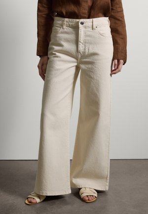 Person trägt cremefarbene, weit geschnittene Hosen, braunes Langarmoberteil und cremefarbene Sandalen, steht auf grauem Boden vor einer weißen Wand.