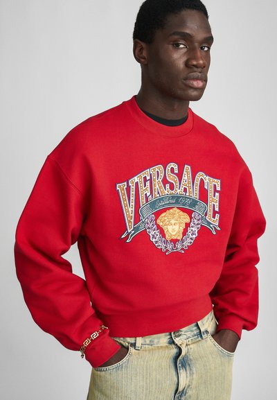 Roter Sweatshirt mit "VERSACE"-Logo und Grafik eines Kopfes, oversize Passform, gerippte Bündchen und Saum, kombiniert mit hellblauen Jeans.