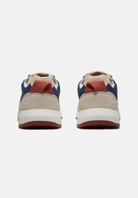 Två sneakers med en beige mocka och marinblå tygöverkropp, röda mesh-heels accentuerande och en vit gummisula. Bakifrån med snörning och sömnad.