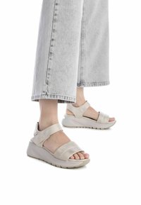 Las sandalias beige con cuña cuentan con correas perforadas, correas ajustables en el tobillo y una suela flexible. El calzado se combina con jeans de pierna ancha de color gris claro.