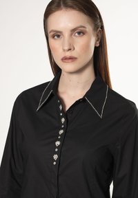 Camicia nera con colletto strutturato, decorata con strass argentati lungo il colletto e la patta dei bottoni. Tessuto a trama liscia.