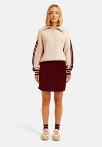 Pull en tricot crème avec un demi-zip, associé à une jupe en velours côtelé bordeaux avec deux poches avant. Baskets blanches et chaussettes rayées.