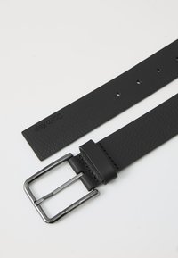 Zwarte lederen riem met een gestructureerd oppervlak, zilveren metalen gesp en vijf verstelbare gaten. Logo in reliëf nabij het uiteinde.
