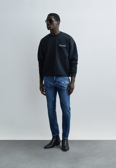 Άνδρας που φοράει μαύρα γυαλιά ηλίου, μαύρο φούτερ με λευκό λογότυπο, μπλε τζιν με φθορές και μαύρα μποτάκια, στέκεται μπροστά σε απλό γκρι τοίχο.