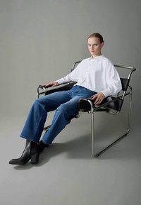 Črne gležnjarje na peto, široke jeans hlače in bela srajca na gumbe nosi oseba, ki sedi na minimalistični kovinski in usnjeni stolu.