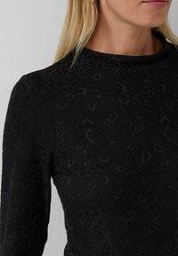 Vrouw in een zwart shirt met lange mouwen en subtiele panterprintstructuur, met een hoge hals, zichtbaar is een deel van het gezicht en het blonde haar.