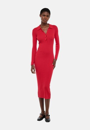 Femme portant une robe midi rouge côtelée ajustée à manches longues, avec col et boutons, associée à des ballerines Mary Jane noires, debout sur fond blanc.