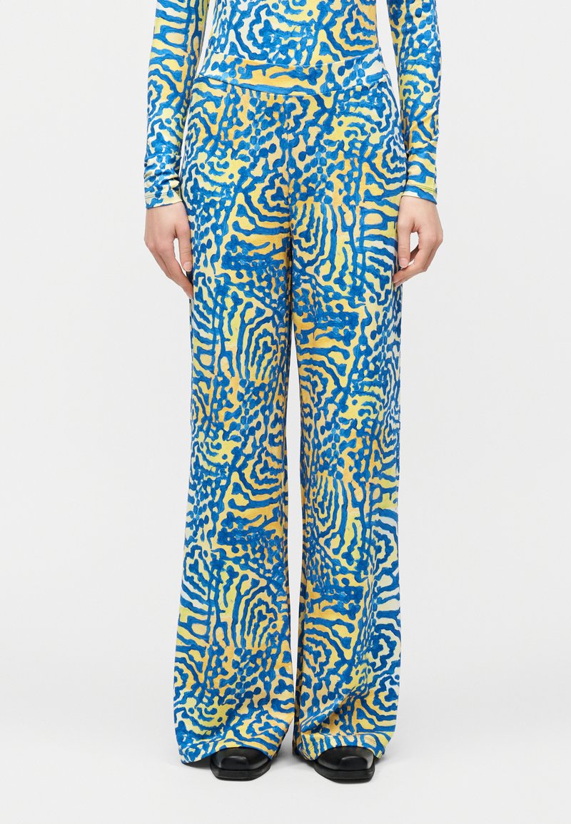 Παντελόνια με φαρδύ πόδι και αφηρημένα μπλε και κίτρινα στροβιλίσματα, φορεμένα με αντίστοιχο μακρυμάνικο τοπ και μαύρες παπούτσια με τετράγωνη μύτη.