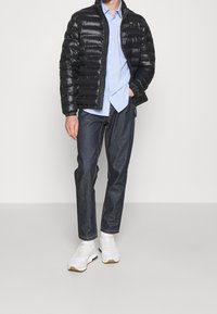 Giacca a vento nera, leggera e con una texture lucida, indossata sopra una camicia azzurro chiaro, abbinata a jeans di jeans scuri e sneakers bianche.