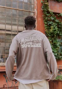 Homme portant une chemise à manches longues beige avec le texte « Mountains Nowhere » et un graphisme de voiture dans le dos, debout devant une fenêtre rouillée et un mur recouvert de lierre.