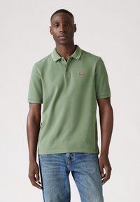 Green polo shirt with a two-button placket, white collar trim, and red logo patch; paired with blue jeans. Smooth texture, casual design.
