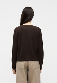 Γυναίκα με μακριά σκούρα μαλλιά που φοράει σκούρο καφέ πουλόβερ με μακριά μανίκια και χαλαρά μπεζ παντελόνια, γυρισμένη με την πλάτη προς ένα απλό φόντο.