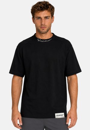 Mann mit kurzem lockigem Haar trägt ein schlichtes schwarzes oversized T-Shirt mit weißem Text am Kragen und einem Aufnäher nahe dem Saum, steht vor einfachem Hintergrund.