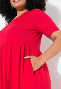 Robe rouge à manches courtes, décolleté en V et jupe froncée. Présente un détail de boutons sur le devant et des poches latérales. Tissu lisse et extensible.