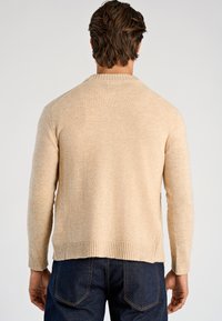 Ljusbeige stickad tröja med ribbad fåll och muddar, med rund halsringning och raglanärmar. Bärs med mörka jeans, sedd bakifrån.