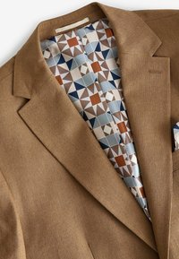 Brauner Blazer aus strukturierter Stoff mit einem gefütterten Innenleben im geometrischen Muster in gedämpften Farben, darunter Blau, Beige und Rost.