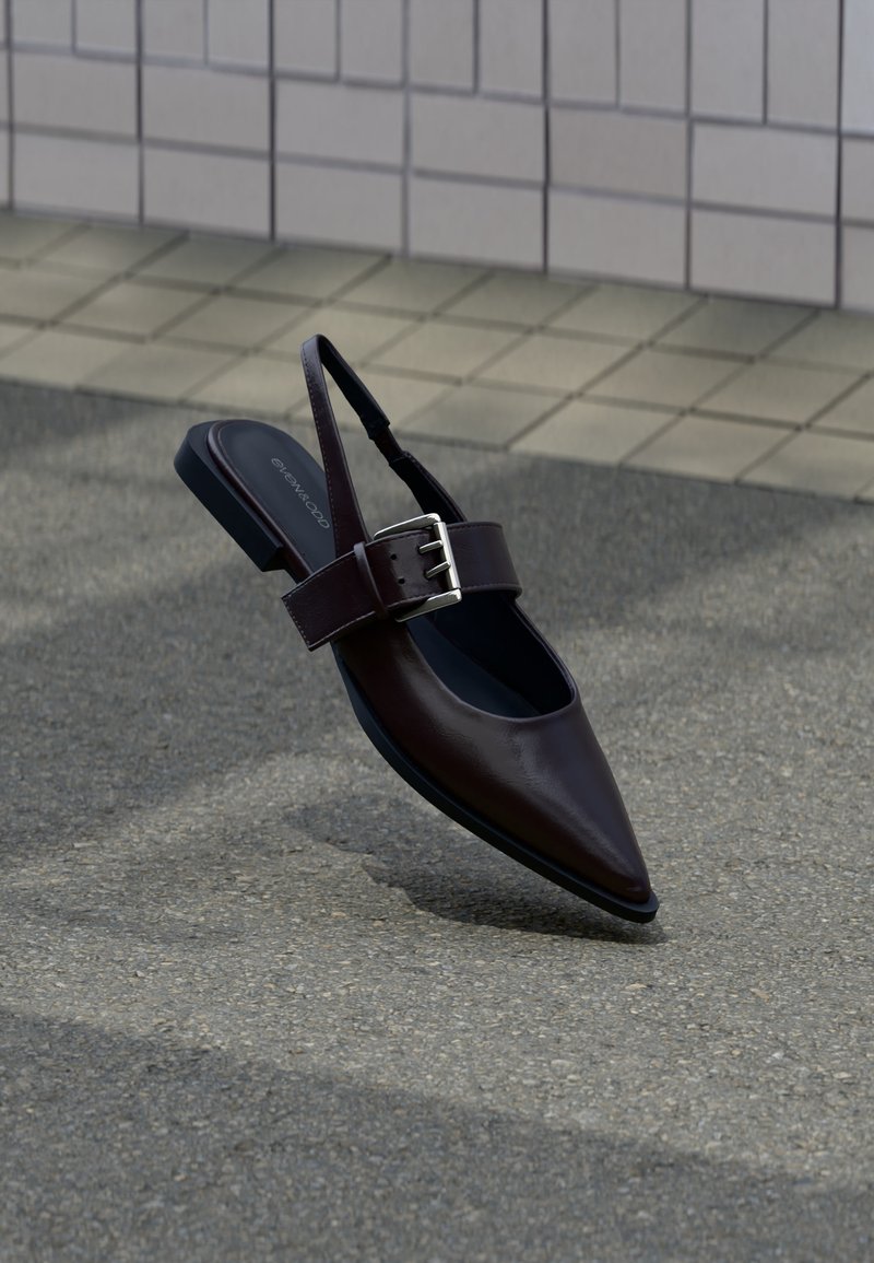 Chaussure plate à bout pointu marron foncé avec bride arrière, large sangle et grande boucle argentée sur l'asphalte près d'un mur carrelé.
