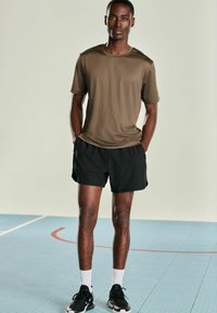 Kortærmet, olivengrøn sportstrøje; kombineret med sorte shorts. Modellen står på et tekstureret blåt gulv, iført sorte sneakers og hvide sokker.
