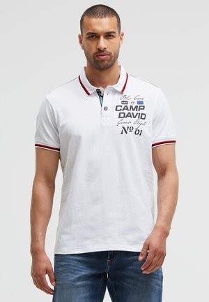 Mann trägt weißes Poloshirt mit rotem und schwarzem Besatz und blaue Jeans, steht vor schlichtem hellem Hintergrund.