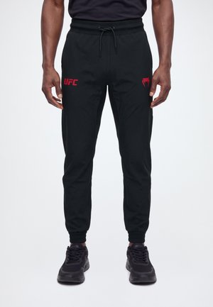 Vista frontal de un modelo con pantalones de chándal negros de UFC con logotipos rojos, cintura con cordón y zapatillas negras.