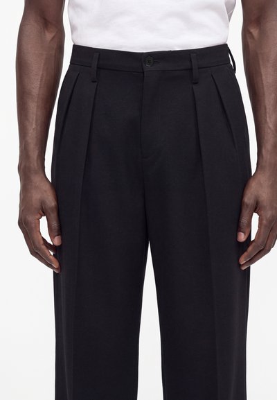 Mann trägt schwarze, plissierte Hose mit hohem Bund und ein weißes, in die Hose gestecktes Hemd, die Hände entspannt an den Seiten, vor weißem Hintergrund.