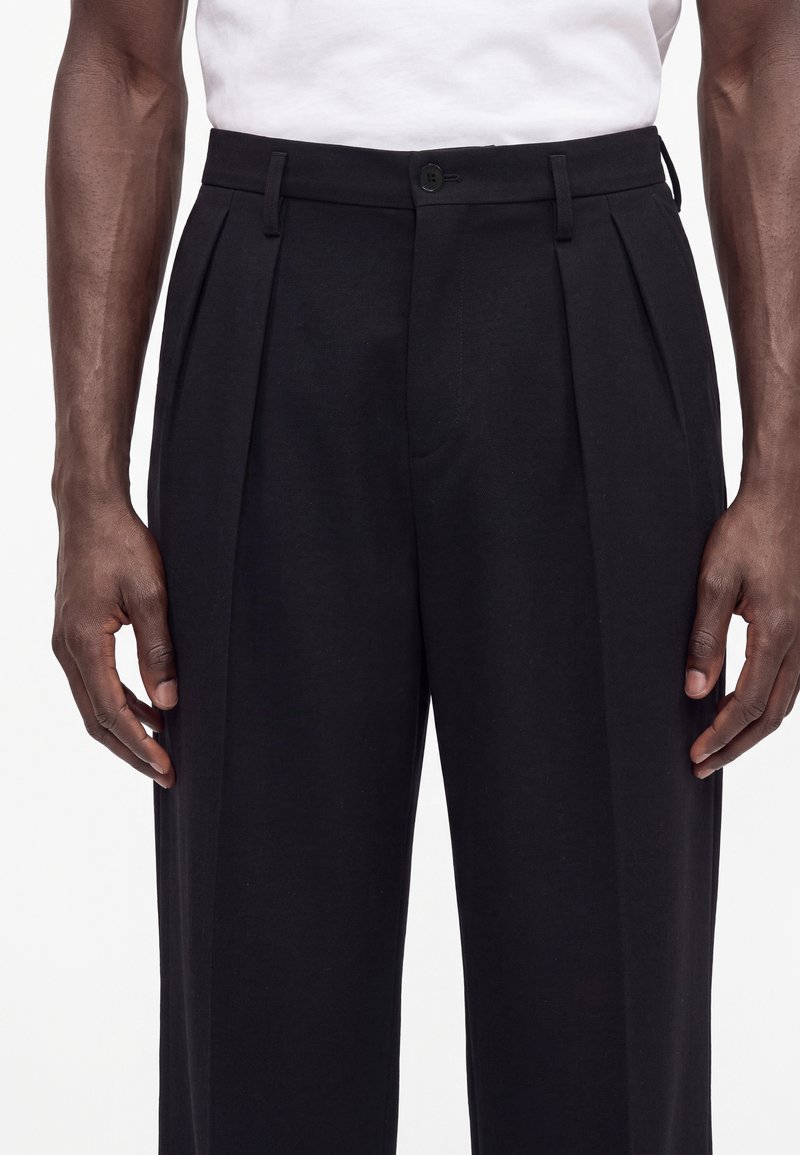 Hombre con pantalones negros plisados de cintura alta y camisa blanca metida, con las manos relajadas a los lados, sobre un fondo blanco.
