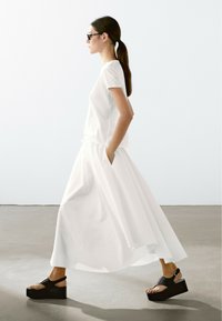 Witte katoenen t-shirt gecombineerd met een luchtige witte rok met zijzakken. Model draagt zwarte platform sandalen met een band en gesp.