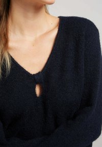 Pull en laine mélangée bleu marine avec un décolleté en V et un petit détail en forme de clé au niveau de la poitrine. Texture douce, coupe décontractée et manches longues.