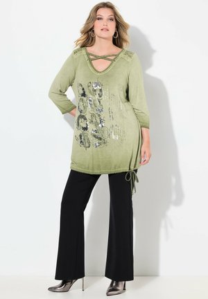 Frau trägt eine olivgrüne Langarm-Tunika mit glitzerndem Text, schwarze Schlaghosen und silberne High-Heels-Stiefel und steht vor einem weißen Hintergrund.