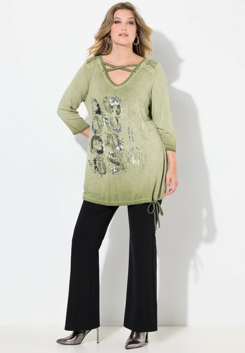 Frau trägt eine olivgrüne Langarm-Tunika mit glitzerndem Text, schwarze Schlaghosen und silberne High-Heels-Stiefel und steht vor einem weißen Hintergrund.