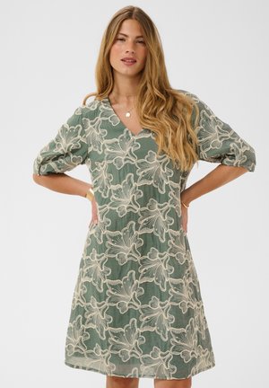 Femme aux longs cheveux blonds portant une robe verte avec un motif en dentelle florale blanche, debout les mains sur les hanches devant un fond uni.