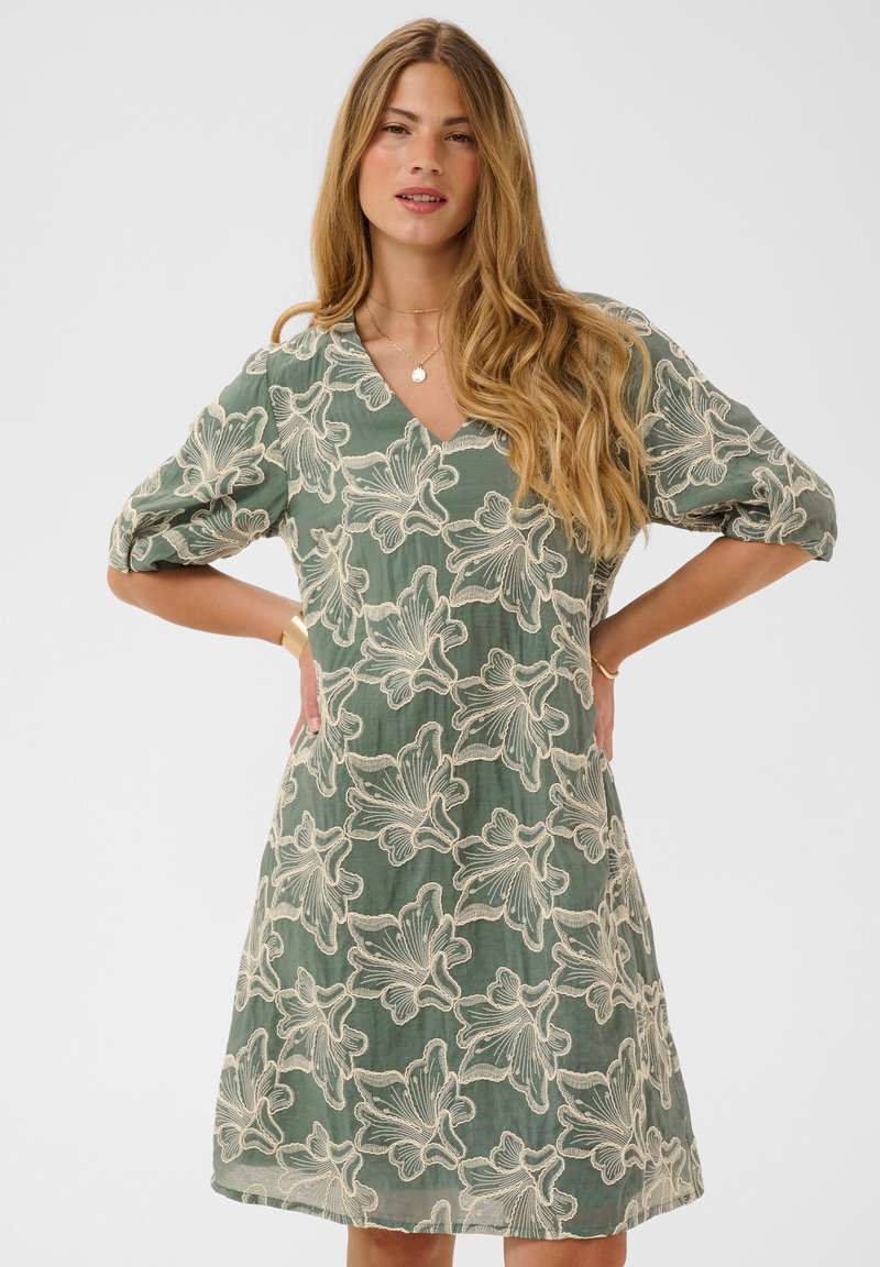 Femme aux longs cheveux blonds portant une robe verte avec un motif en dentelle florale blanche, debout les mains sur les hanches devant un fond uni.