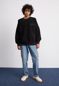 Sweat-shirt en polaire noire avec une finition texturée et un logo brodé, associé à un pantalon cargo en denim clair avec plusieurs poches.