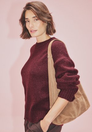 Suéter de punto burdeos con cuello redondo y mangas enrolladas, acompañado de un bolso de ante color beige. Textura suave sobre un fondo rosa.