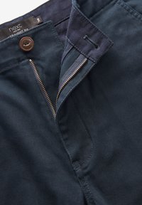 Marineblaue Hose mit Reißverschluss und goldfarbenem Reißverschluss, braunem Knopfverschluss und Seitentaschen, aus einem glatten Stoff gefertigt.