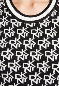 Gros plan d'un haut en maille noire avec un motif répétitif du logo blanc DKNY et un col rond côtelé blanc et noir.