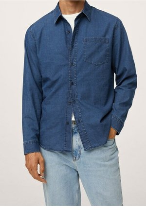 Mann trägt ein dunkelblaues Jeanshemd mit Knöpfen über einem weißen T-Shirt und hellblauen Jeans, eine Hand in der Tasche, vor schlichtem Hintergrund.