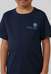 Marineblauwe katoenen T-shirt met korte mouwen. Voorzien van een grafische print aan de voorkant met de tekst "ONLY THE" en een cirkelvormig ontwerp. Eenvoudige en stevige textuur.