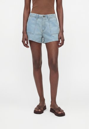 Persoană îmbrăcată în pantaloni scurți din denim bleu deschis, top maro fără mâneci și sandale maro cu platformă, stând în fața unui fundal alb.