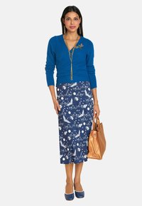 Blauer Strickpullover mit V-Ausschnitt, gemusterter Midirock in Dunkelblau mit weißen Designs, goldene Handtasche und blaue Absätze. Lange Kette und Ohrringe.