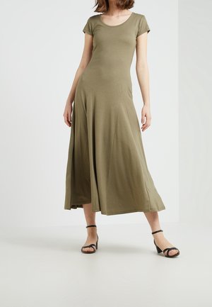 Robe maxi olive verte à manches courtes avec un col rond, jupe fluide et texture lisse. Portée avec des sandales à talons noirs.