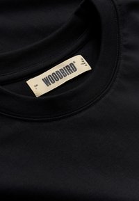 Κοντινό πλάνο στο γιακά μαύρου μπλουζιού crewneck με ετικέτα της μάρκας Woodbird σε εκρού χρώμα που δείχνει το μέγεθος και τις λεπτομέρειες μετρήσεων.